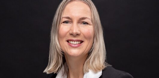 Professional headshot of Léonie Koning, wearing a black blazer and white blouse, smiling against a dark background.